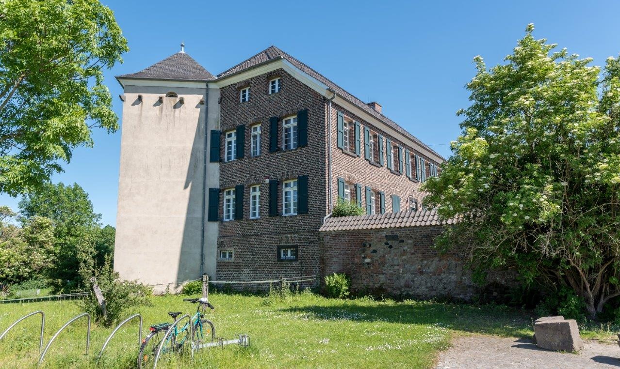 Der hell verputzte Turm von Haus Bürgel und seine anschließende Ziegelmauer mit antiken Resten vor blauem Himmel. Im Vordergrund befindet sich eine Grünfläche und ein Fahrradständer mit Fahrrad.