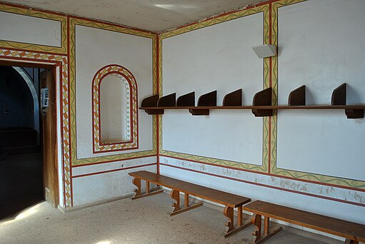 Foto des Apodyteriums (Umkleideraums) der rekonstruierten Thermen der römischen Herberge im LVR-Archäologischen Park Xanten. Zu sehen sind Holzbänke und ein durchgehendes hölzernes Wandbord mit Querteilungen. Die weißen Wände des Raums, eine halbrunde Wandnische und die Türöffnung sind mit unterschiedlich gemusterten Ornamentstreifen in den Farben Gelb, Grün, Lehm und Ziegelrot umrahmt.
