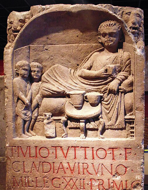 Grabstein mit Reliefdarstellung für den Legionär Titus Iulius Tuttius, der auf einem Speisesofa liegt. Am Fußende dieses Sofas stehen zwei kleinere Sklaven in offensichtlich demütiger Haltung. Der Größenunterschied ist symbolisch gemeint und drückt den ungleichen gesellschaftliche Rang zwischen ihnen und ihrem Eigentümer aus. Römisch-Germanisches Museum Köln.