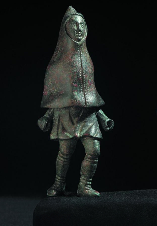 Fotografie einer Bronzestatuette eines (Land)arbeiters in wetterfester Kleidung: Über die Tunika hat er einen Kapuzenmantel gezogen. Die Wickelgamaschen sind mit Bändern oberhalb der Knöchel und unterhalb der Knie befestigt. Rheinisches Landesmuseum Trier.