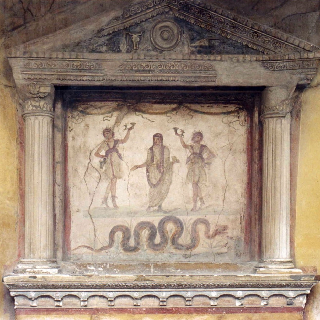 Foto eines römischen Hausaltars aus dem Haus der Vettier in Pompeji, vor 79 n. Chr. Der Altar ist wie ein Miniaturtempel aufgebaut. Das von jeweils einer Säule mit korinthischem Kapitell gerahmte Bildfeld in der Mitte zeigt eine Malerei von drei Gottheiten und einer Schlange. Der Altar ist mit einem Dreiecksgiebel überdacht und leicht in die Hauswand eingenischt.
