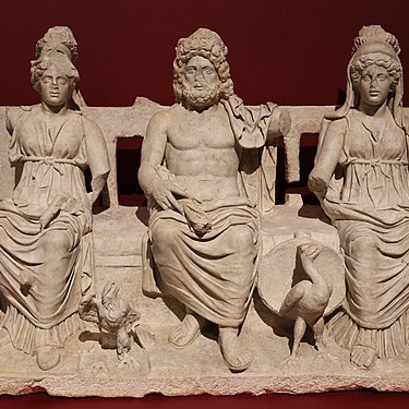 Steinerne Tafel mit Relief der Kapitolinischen Trias aus dem 2. Jh. n. Chr.. Von links nach rechts: Minerva (Helm, Eule) – Jupiter (Mantel, Adler) - Juno (Schleier, Pfau). Übliche weitere Attribute der Gottheiten wie Blitz und Zepter sind nicht erhalten. Museo civico archeologico, Guidonia Montecelio.
