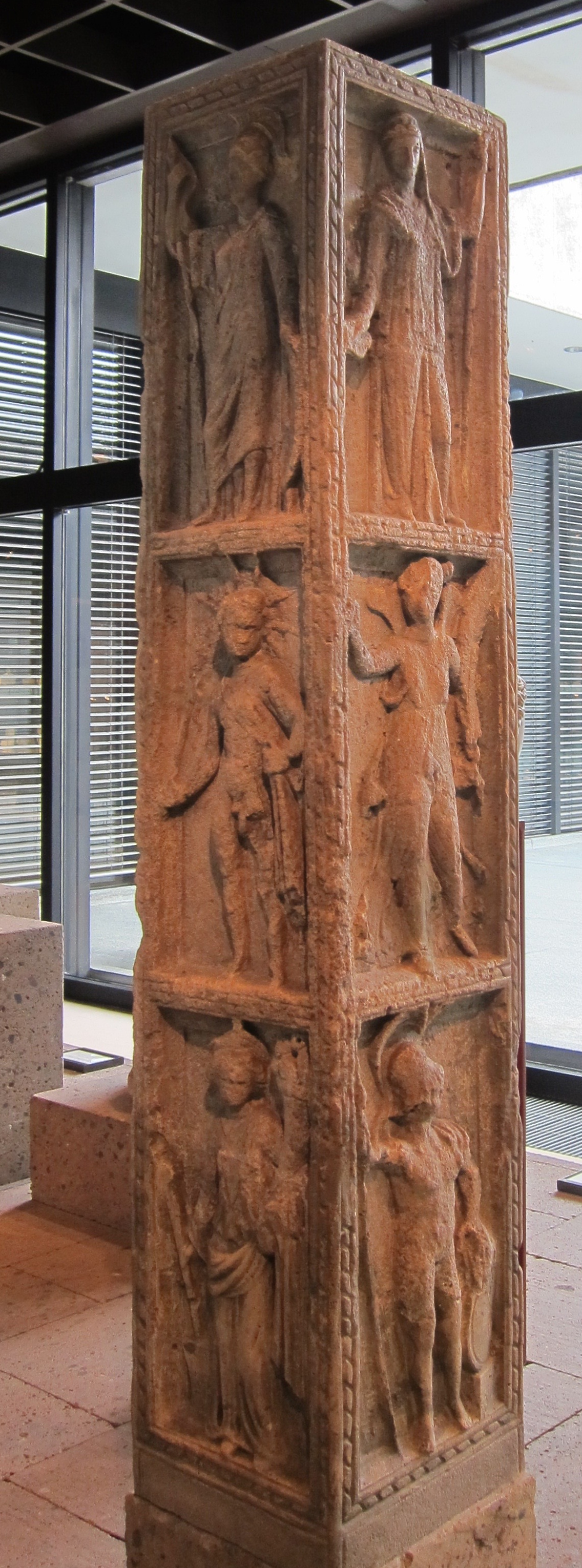 Foto eines steinernen Jupiter-Pfeilers mit Götterdarstellungen auf drei Seiten. Rechts: Juno (Schleier, Schale) - Viktoria (Flügel, Palmrispe) - Mars (Helm, Lanze Schild). Mitte: Minerva (Kleid, Helm, Lanze, Schild) - Sol (Strahlenkrone) - Fortuna (Füllhorn, Steuerruder). Links:  Ceres (Ähre) - Vulkan (Kappe, Handwerkertunika) - Venus (Spiegel). Römisch-Germanisches Museum Köln.