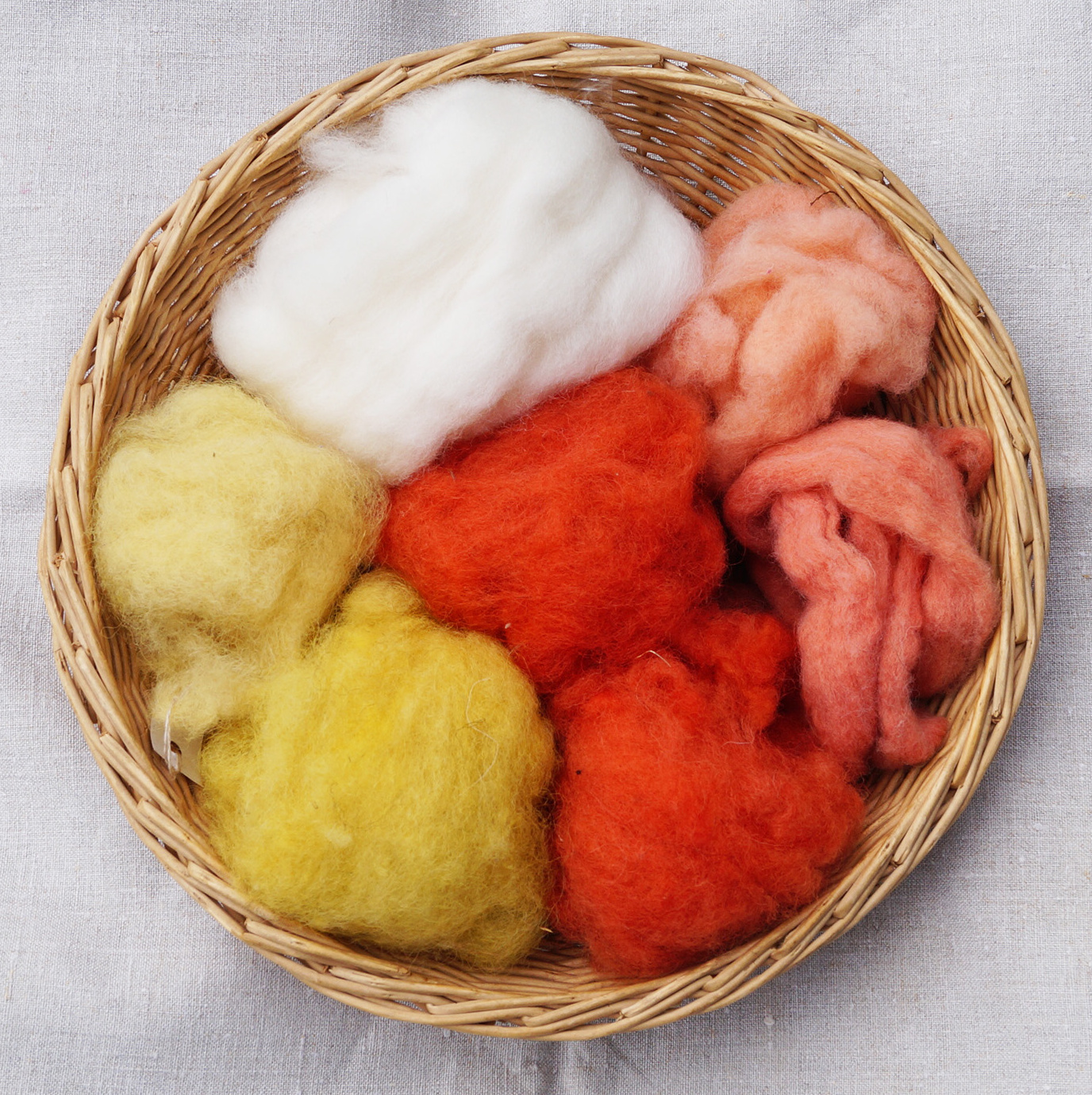 Foto eines geflochtenen Körbchens mit unterschiedlichen Büscheln gewaschener Schafswolle: ungefärbt (naturweiß) bzw. gefärbt mit Krappwurzel (rot/rosa), Resede (leuchtend gelb) und Färberginster (hellgelb).